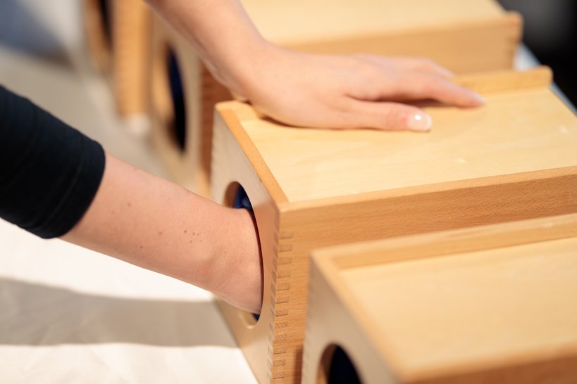 Eine Person hat ihre Hand in einer Holzbox, in der verschiedene Dinge ertastet werden können.