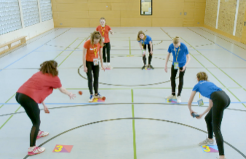 Ein Foto einer Spielszene des Spiels 'FAIR WERFEN'. Ein rotes Team spielt gegen ein blaues Team in einer Turnhalle.