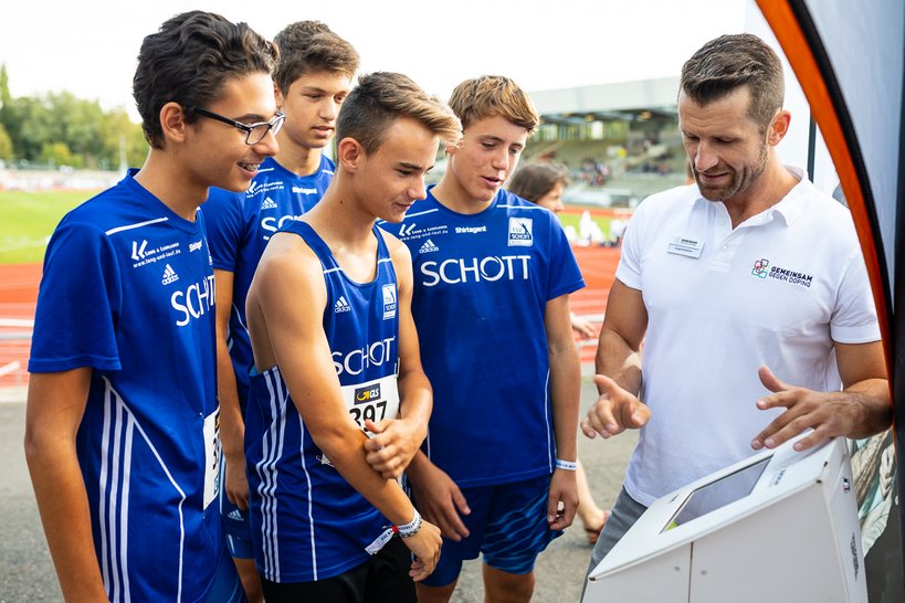 Ein Mitarbeiter von 'GEMEINSAM GEGEN DOPING' erklärt jungen Sportlern etwas an einem Display.